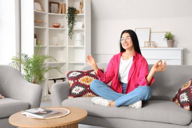 Evdeki kanepede meditasyon yapan genç bir kadın.