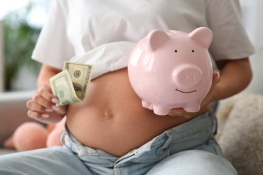 Kumbarası ve parası olan hamile bir kadın evdeki kanepede oturuyor, yakın plan. Annelik Faydası konsepti