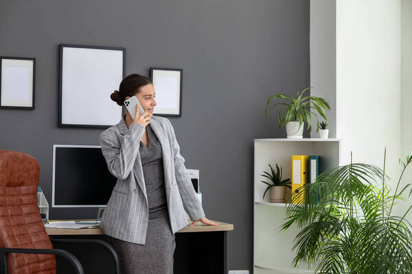Работница офиса говорит по мобильному телефону возле кресла и рабочего места в стильном офисе