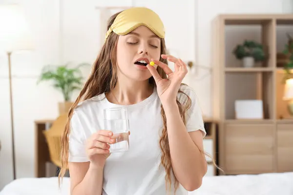 stock image Young woman with glass of water taking sleeping pill in bedroom