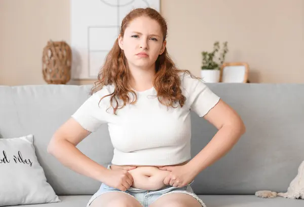 stock image Sad young woman in tight shorts sitting on sofa at home. Weight gain concept