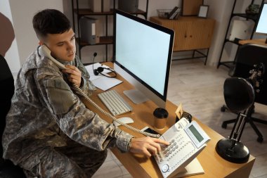 Genç erkek asker merkezde telefonla konuşurken bilgisayarla çalışıyor.