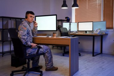 Kulaklığı olan genç bir erkek asker geceleyin merkezde bilgisayar kullanıyor.