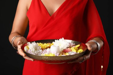 Beautiful young Indian woman in sari holding plate with candles and flowers on black background, closeup. Divaly celebration clipart