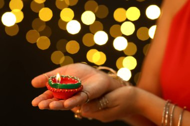 Beautiful young Indian woman in traditional sari with diya lamp against blurred lights on black background, closeup. Divaly celebration clipart