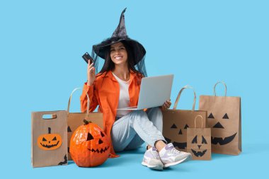 Cadı şapkalı, laptoplu ve kredi kartlı genç bir kadın Cadılar Bayramı için mavi arka planda online alışveriş yapıyor.
