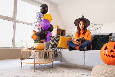 Cadı şapkalı genç Asyalı kadın büyü kitabıyla kanepede oturuyor ve evde Cadılar Bayramı balonları var.