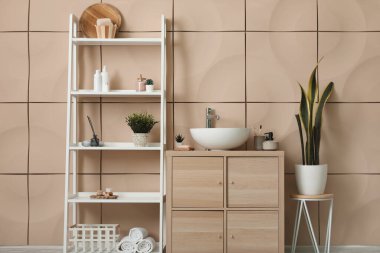 Lavabo kasesi, ahşap dolap ve bej duvarın yanında raf ünitesi olan banyonun içi.