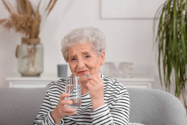 Evde bir bardak su ve hap taşıyan yaşlı bir kadın var.