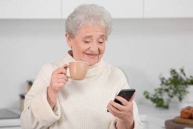 Mutfakta cep telefonu kullanan çaylı yaşlı bir kadın.