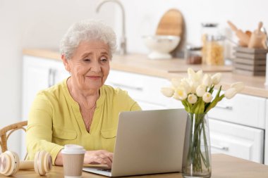 Mutfakta dizüstü bilgisayar kullanan yaşlı bir kadın.