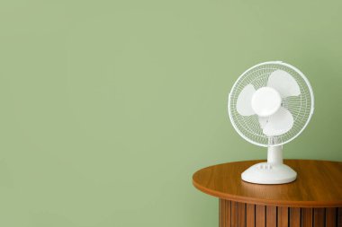 Yeşil duvarın yanında modern elektrikli vantilatörü olan ahşap bir sehpa.