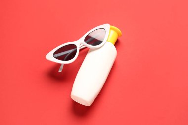 Kırmızı arka planda güneş kremi şişesi olan güneş gözlüğü.