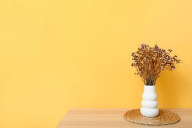 Kurumuş çiçekli vazo ve sarı duvarın yanındaki sehpanın üzerinde paspas.