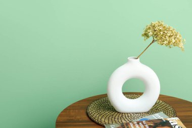 Yeşil duvarın yanındaki sehpanın üzerinde kurutulmuş ortanca vazosu, dergi ve paspas var.