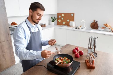 Genç adam mutfakta kızarmış sebzelere baharat katıyor.