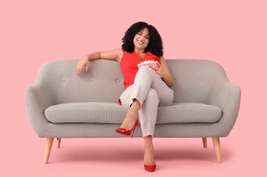 Güzel bir kadın elinde patlamış mısırla kanepede pembe arka planda film izliyor.