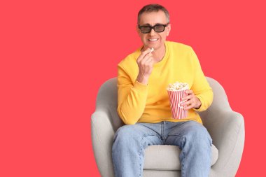 3 boyutlu gözlüklü ve patlamış mısırlı olgun bir adam kırmızı arka planda koltukta film izliyor.