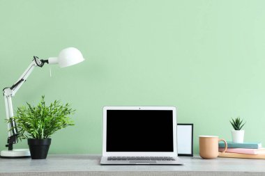Ofiste yeşil duvarın yanında modern dizüstü bilgisayarı, lambası ve bitkisi olan şık bir iş yeri.