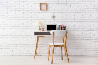 Ofisteki beyaz tuğla duvarın yanında modern dizüstü bilgisayarı, sandalyesi ve lambası olan şık bir iş yeri