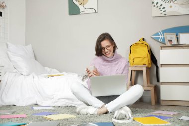 Bardaklı ve dizüstü bilgisayarlı güzel öğrenci yatak odasında çalışıyor.