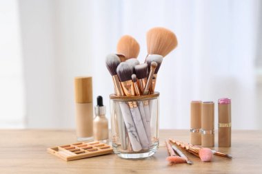 Makyaj fırçaları ve evdeki ahşap masada farklı dekoratif kozmetik ürünleri olan tutucu.