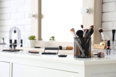 Makyaj fırçaları ve farklı dekoratif kozmetik ürünleri olan tutucu evdeki beyaz masada.