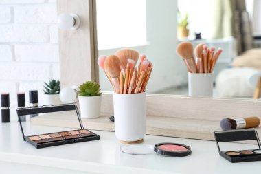 Makyaj fırçaları ve farklı dekoratif kozmetik ürünleri olan tutucu evdeki beyaz masada.