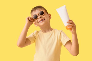 Sarı arka planda güneş kremi sürmüş, güneş gözlüklü şirin bir çocuk.