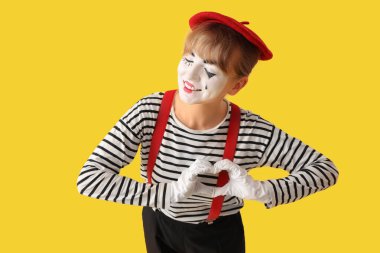 Kadın pandomimci elleri sarı arka planda kalp yapıyor.