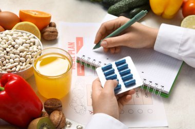 Kadın beslenme uzmanı deftere yazıyor ve sağlıklı yiyeceklerin yanında haplar tutuyor.