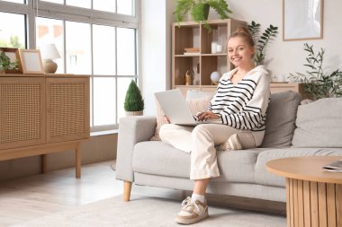 Evdeki koltukta modern laptop kullanan genç bir kadın.