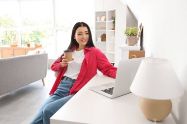 Evdeki masada dizüstü bilgisayar kullanan bir fincan kahveli genç kadın.