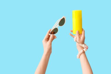 Mavi arka planda güneş kremi, güneş gözlüğü ve kolye şişesi olan kadın elleri.