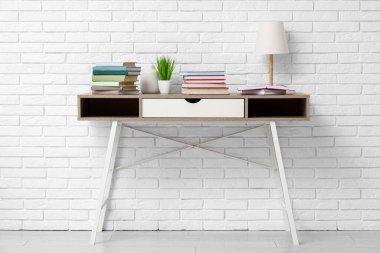 Beyaz tuğla duvarın yanındaki masada yığınla kitap, lamba ve çiçek var.