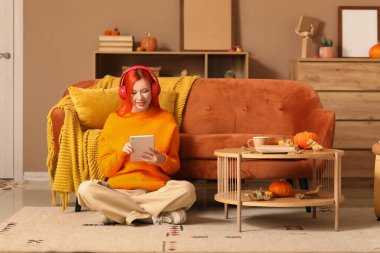 Güz dekoru ve tabletli kulaklıklı güzel, mutlu bir kadın evde yerde oturuyor.