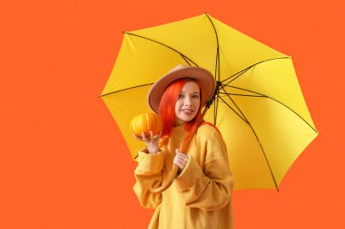 Sonbahar kıyafetleri içinde, turuncu arka planda balkabağı ve şemsiyesi olan genç ve güzel bir kadın.