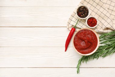Bir kase kırmızı biber sosu ve beyaz ahşap arka planda farklı baharatlar.
