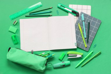 Kalem kutulu boş defter ve yeşil arka planda farklı kırtasiye malzemeleri.