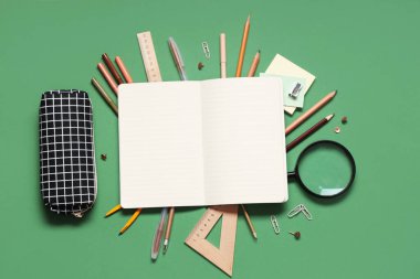 Kalem kutulu boş defter ve yeşil arka planda farklı kırtasiye malzemeleri.