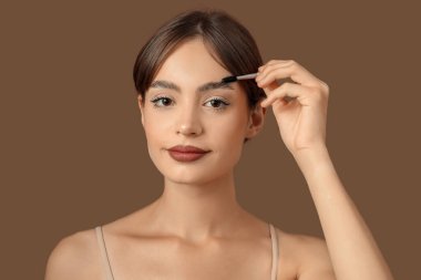 Kahverengi arka planda kaşlarını düzelten genç bir kadın.