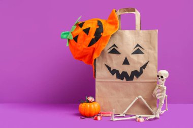 Mor arka planda Cadılar Bayramı için kağıt alışveriş torbası, süsleme ve tatlı kompozisyonu