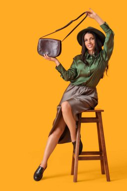 Sonbahar kıyafetli, şık çantalı, sarı arka planda sandalyede oturan güzel, genç ve mutlu bir kadın.