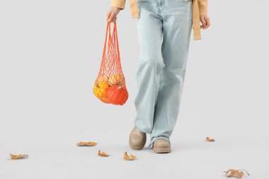 Gri arka planda bir sürü balkabağı ve sonbahar yapraklarıyla dolu sicim çantasıyla güzel genç bir kadın. Yakın plan.