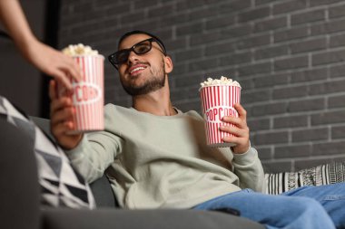 3 boyutlu gözlüklü genç adam akşamları kanepede film izlerken patlamış mısır alıyor.