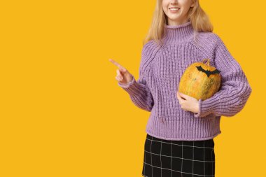 Cadılar Bayramı balkabağı ve kağıt yarasayla güzel, genç ve mutlu bir kadın sarı arka plandaki bir şeyi işaret ediyor. Yakın plan.