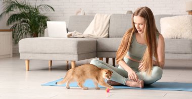 Evde yoga minderinde kedi yavrusuyla oynayan genç bir kadın.