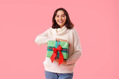 Pembe arka planda Noel hediyesi olan kalın kazaklı genç bir kadın.