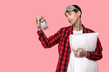 Pembe arka planda yastıklı ve çalar saati olan pijamalı dövmeli genç adam.