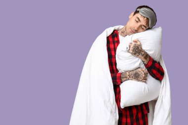 Mor arka planda yastıklı ve battaniyeli pijamalı dövmeli genç adam.